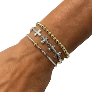 May include: Four bracelets are stacked on a wrist. One bracelet features silver crosses, another has small silver beads, and two have gold beads. The bracelets are a mix of silver and gold tones.