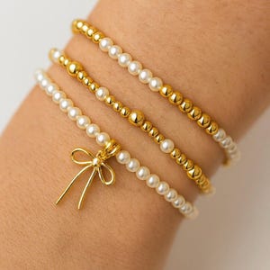 Puede incluir: Se muestran cuatro pulseras de oro y perlas. Tres pulseras están hechas de pequeñas cuentas doradas y perlas blancas. La cuarta pulsera tiene un dije de lazo dorado. Las pulseras están en una muñeca.