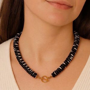 May include: A black and white beaded necklace with a gold-colored clasp. The necklace features alternating black and white circular beads. The clasp is a simple, circular design. The necklace is worn around the neck.