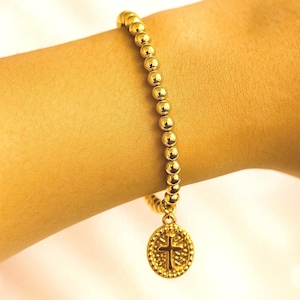 May include: A gold-coloured beaded bracelet with a cross pendant. The bracelet is made of small, round beads and a circular pendant with a detailed cross design. The bracelet is worn on a wrist, showing its shiny, golden appearance.