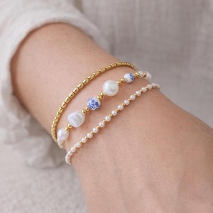 May include: Three bracelets stacked on a wrist. The top bracelet is gold-colored with small rectangular beads. The middle bracelet has white pearls and blue and white ceramic beads. The bottom bracelet is made of small white pearls and gold-colored beads.