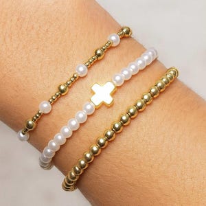 Puede incluir: Tres pulseras de cuentas en oro y blanco. Una pulsera presenta cuentas doradas, otra perlas blancas, y la tercera una mezcla de cuentas doradas y perlas blancas. La pulsera central tiene un dije de cruz dorada.