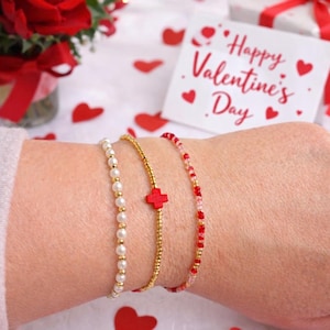May include: Three bracelets on a wrist. One is pearl, one is gold with a red cross, and one is red and pink beads. A Valentine's Day card with hearts and the words "Happy Valentine's Day" is in the background.
