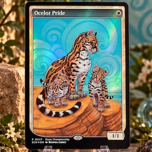 Puede incluir: Una carta coleccionable rectangular titulada Ocelot Pride con una ilustración holográfica de un ocelote adulto y dos crías sobre una roca frente a un cielo azul con nubes arremolinadas, borde negro y recuadros de texto blancos.