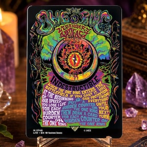 May include: A rectangular trading card featuring a black background with vibrant, psychedelic-style artwork. The text reads The One Ring, Legendary Artifact, Indestructible, and includes game rules. The card displays a central flaming eye motif.