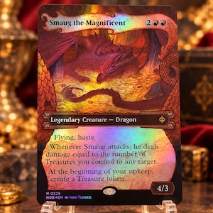 Puede incluir: Una carta coleccionable holográfica titulada Smaug the Magnificent que muestra un dragón rojo sobre monedas de oro. El texto incluye Flying, haste, habilidades relacionadas con fichas Treasure y una fuerza y resistencia de 4/3.