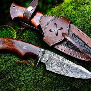 May include: A handcrafted hunting knife with a patterned steel blade and a dark wood handle. The knife is displayed with a brown leather sheath, set against a backdrop of green moss. The blade has a unique wave-like pattern.