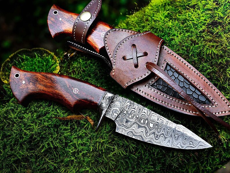 May include: A handcrafted hunting knife with a patterned steel blade and a dark wood handle. The knife is displayed with a brown leather sheath, set against a backdrop of green moss. The blade has a unique wave-like pattern.