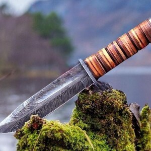May include: A handcrafted hunting knife with a patterned steel blade and a layered wooden handle. The knife rests on a moss-covered surface, with a blurred natural background. The blade has a unique, textured pattern.