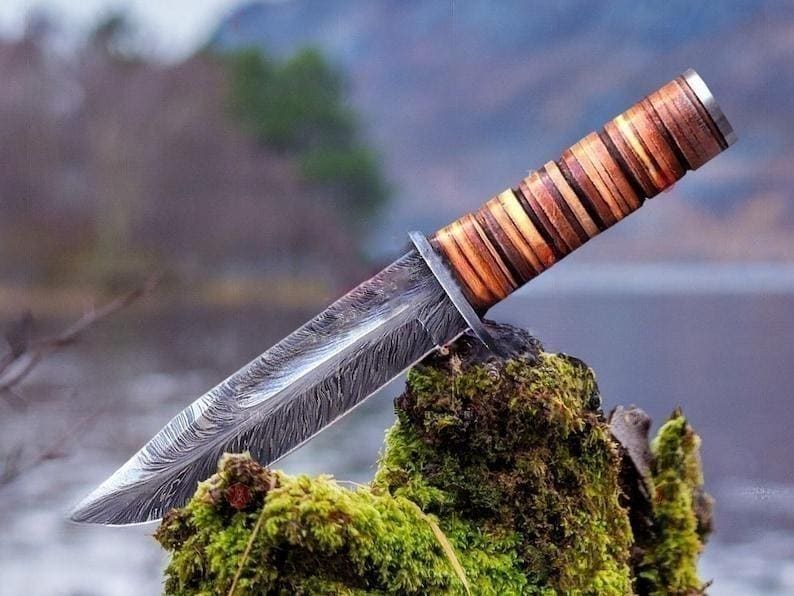May include: A handcrafted hunting knife with a patterned steel blade and a layered wooden handle. The knife rests on a moss-covered surface, with a blurred natural background. The blade has a unique, textured pattern.