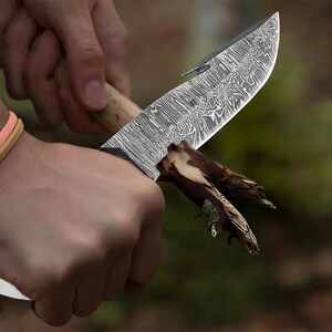 May include: A Damascus steel knife with a wooden handle is held in a hand, cutting a small piece of wood. The blade has a curved shape with a hook at the tip. The knife's surface has a unique, swirling pattern.