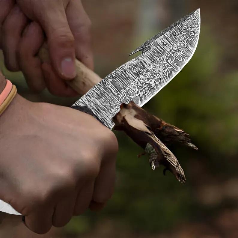 May include: A Damascus steel knife with a wooden handle is held in a hand, cutting a small piece of wood. The blade has a curved shape with a hook at the tip. The knife's surface has a unique, swirling pattern.