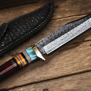 May include: A handcrafted hunting knife with a patterned blade and a handle featuring blue, orange, and brown accents. The knife rests on a wooden surface next to a black leather sheath.