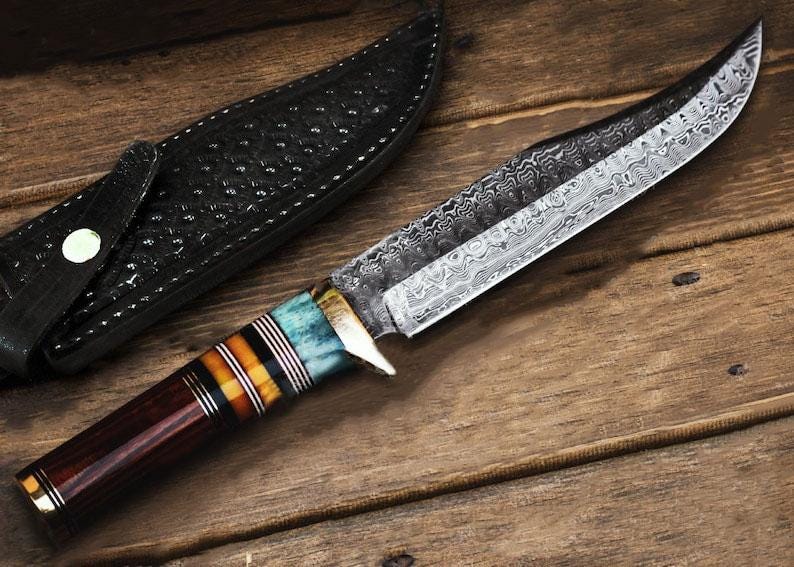 May include: A handcrafted hunting knife with a patterned blade and a handle featuring blue, orange, and brown accents. The knife rests on a wooden surface next to a black leather sheath.