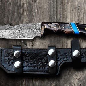 May include: A Damascus steel knife with a patterned blade and a handle featuring wood, black, and blue accents. The knife rests above a black leather sheath with silver rivets, all set against a wooden background.