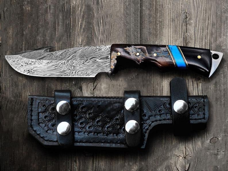 May include: A Damascus steel knife with a patterned blade and a handle featuring wood, black, and blue accents. The knife rests above a black leather sheath with silver rivets, all set against a wooden background.