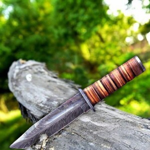May include: A handcrafted knife with a textured blade and a layered leather handle. The blade has a dark, patterned finish, and the handle is wrapped in shades of brown. The knife rests on a weathered wooden surface against a blurred green background.