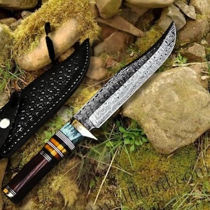 May include: A Damascus steel hunting knife with a patterned blade and a multi-coloured handle. The handle features brown, orange, and blue accents. A black leather sheath is visible next to the knife. The knife is displayed on moss and rocks.