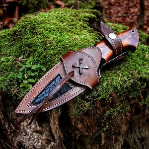 May include: A handcrafted hunting knife with a wooden handle and a leather sheath. The sheath is brown with a cross design and white stitching. The knife is resting on a bed of green moss.
