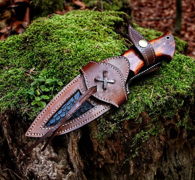 May include: A handcrafted hunting knife with a wooden handle and a leather sheath. The sheath is brown with a cross design and white stitching. The knife is resting on a bed of green moss.