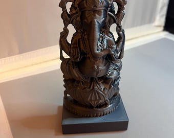 Puede incluir: Una estatua negra, detallada, impresa en 3D de la deidad hindú Ganesha. La estatua está sentada sobre una base cuadrada.