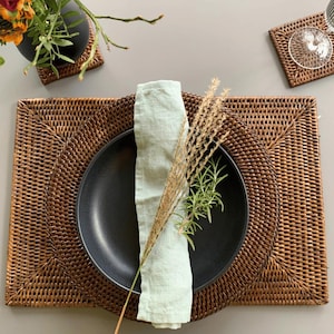 May include: A brown woven placemat with a black plate and a light green linen napkin. The placemat has a square shape with rounded corners. The plate is round and has a slightly raised edge. The napkin is folded in half and placed on top of the plate. There are dried grasses and rosemary sprigs on top of the napkin.