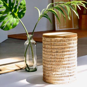 Puede incluir: Una cesta cilíndrica tejida con una tapa de madera. La cesta está hecha de material tejido de color marrón claro. A la izquierda de la cesta hay una botella de vidrio transparente con tallos y hojas verdes. Una hoja verde grande está en la botella.