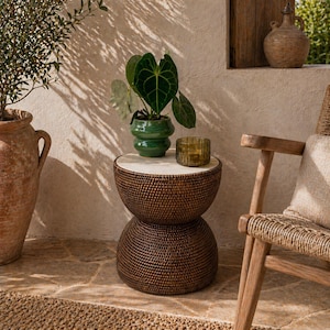 Op de afbeelding: Een bruine geweven bijzettafel met een wit blad, met een groene plant in pot en een klein amberkleurig glas. De tafel is ongeveer 50 cm hoog. Een geweven stoel en een grote terracotta pot zijn ook zichtbaar.