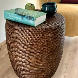 May include: A brown wicker side table with a book titled "Die Dämonen" by Patricia Cornwell on top. A small green ceramic ball and a dark green ceramic bowl are also on the table.