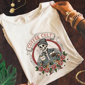Op de afbeelding: Crèmekleurig T-shirt met een afbeelding van een skelet met een koffiekopje en een kan, met de woorden "COFFEE CULT" in een banner. Het ontwerp bevat rozen, dolken en het woord "JAVA" op de beker.