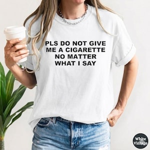 Op de afbeelding: Wit T-shirt met de tekst "PLS DO NOT GIVE ME A CIGARETTE NO MATTER WHAT I SAY" in het zwart. Het shirt wordt gedragen met een blauwe spijkerbroek. Een koffiekopje wordt in de linkerhand gehouden. Het shirt is van White Vintage.