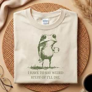 Op de afbeelding: Crèmekleurig T-shirt met een groene illustratie van een kikker die een mok vasthoudt. De kikker heeft grote ogen en een contemplatieve uitdrukking. De tekst hieronder luidt: "I HAVE TO SAY WEIRD STUFF OF I'LL DIE."