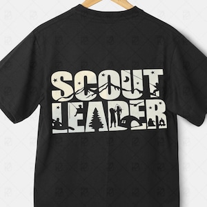 May include: Black t-shirt with the words "SCOUT LEADER" in a white, stylized font. The design incorporates camping and outdoor elements, including a tent, campfire, and silhouettes of people fishing and hiking. A wooden hanger supports the shirt.