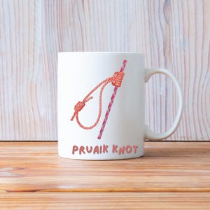 May include: White ceramic mug featuring a graphic of a Pruvaik knot in shades of red and pink. The mug has a curved handle and the text "PRUAIK KNOT" in brown letters.