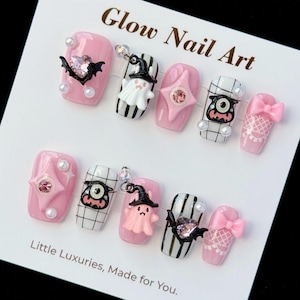 May include: A set of ten press-on nails on a display card labeled Glow Nail Art. The designs feature pink and white colors, black stripes, and grid patterns. Embellishments include 3D ghost figures, bat-winged hearts, pink bows, and rhinestones.