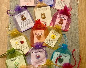 Op de afbeelding: Een verzameling kleine, kleurrijke cadeauzakjes gevuld met kleine cadeautjes en inspirerende boodschappen. De zakjes zijn gemaakt van doorzichtig materiaal en hebben een trekkoord. De boodschappen zijn "You've Got This!" en "A Pocket Hug for My Bestie".