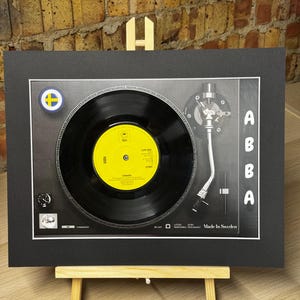 Op de afbeelding: Een ingelijste display met een zwarte vinylplaat op een platenspeler-afbeelding met een geel label in het midden. De tekst ABBA staat verticaal aan de rechterkant. Een klein rond Zweeds vlag-embleem bevindt zich in de linkerbovenhoek. De tekst Made In Sweden staat rechtsonder op de platenspeler-afbeelding.