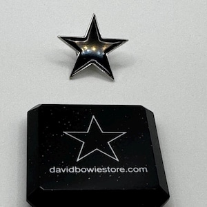 Könnte beinhalten: Ein silberner sternförmiger Ansteckpin liegt über einer schwarzen Box. Der Pin hat eine glänzende, reflektierende Oberfläche. Die schwarze Box hat einen weißen Sternumriss und den Text "davidbowiestore.com" aufgedruckt.