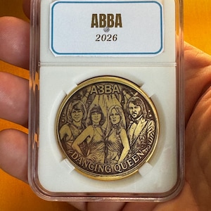 Pode incluir: Uma moeda dourada com a banda ABBA, com o texto "DANCING QUEEN" ao redor da borda. A moeda está dentro de um suporte de plástico transparente com um rótulo branco que diz "ABBA 2026".