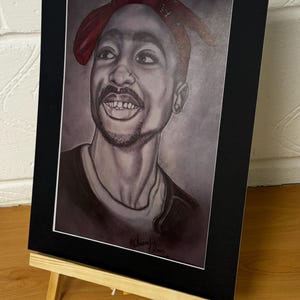May include: A framed black and white portrait of a smiling man wearing a red bandana. The artwork is displayed on a small wooden easel. The portrait has a black mat and is signed in the bottom right corner.