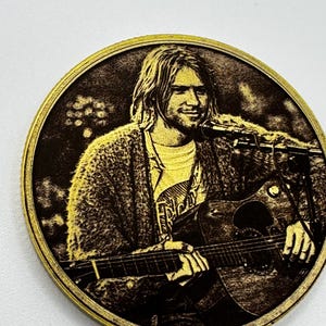 May include: A circular, gold-coloured coin depicting a musician playing a guitar and singing into a microphone. The image is a detailed engraving with a dark brown background and a gold border.