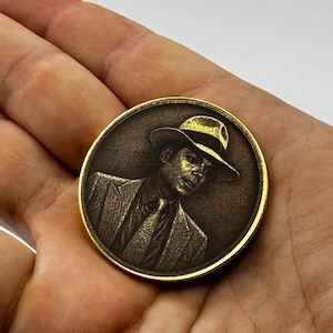 Könnte beinhalten: Eine runde, bronzefarbene Münze mit einem goldfarbenen Rand. Die Münze zeigt ein detailliertes Porträt einer Person mit Hut und Anzug. Das Porträt ist im Vintage-Stil gehalten, mit feinen Linien und Schattierungen.