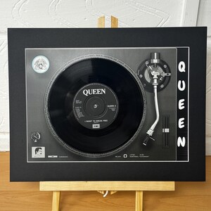 Op de afbeelding: Een ingelijste print met een vintage platenspeler met een Queen-plaat. Het label van de plaat luidt "Queen" en "I Want to Break Free". Het woord "Queen" is verticaal in wit gedrukt aan de rechterkant van het zwarte frame. De print wordt gepresenteerd op een kleine houten ezel.