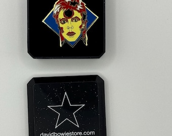 Puede incluir: Un colorido pin de esmalte con un retrato estilizado de una persona con cabello rojo y blanco, piel amarilla y un fondo de diamante azul. El pin está en una caja negra. La caja de abajo tiene un gráfico de estrella y el texto "davidbowiestore.com".