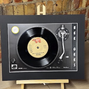 May include: A framed display featuring a black vinyl record centered on a graphic of a turntable. The record label reads Bee Gees, Rest Your Love On Me. The text BEE GEES is printed vertically on the right side, with the word Brothers at the bottom.