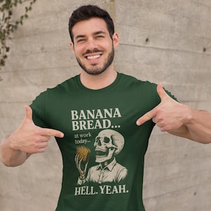 May include: Dark green t-shirt with the text "BANANA BREAD... at work today... HELL. YEAH." A graphic of a skeleton holding banana bread is printed on the shirt. The person is pointing at the shirt.
