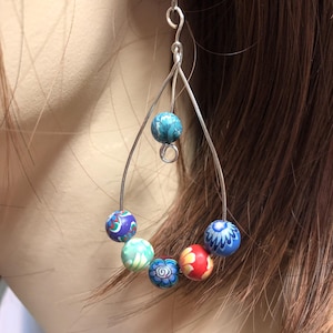 May include: A silver wire earring with a teardrop design. The earring features seven colorful, patterned beads in shades of blue, red, green, and purple. The beads are arranged along the wire, creating a unique and eye-catching accessory.