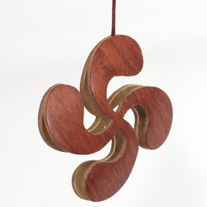 Peut inclure: Un pendentif en bois représentant une croix basque, également appelée Lauburu, suspendu à un cordon marron. Les quatre bras incurvés sont en bois brun-rouge superposé avec un accent en bois plus clair. Le pendentif est sur fond blanc.