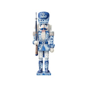 Puede incluir: Un cascanueces decorativo azul y blanco con forma de soldado sosteniendo un rifle. El soldado lleva un sombrero alto y un uniforme con intrincados diseños florales. El cascanueces tiene bigote y barba blancos, y está sobre una base rectangular.