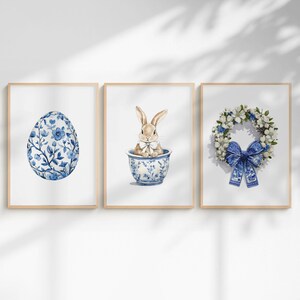 Könnte beinhalten: Drei gerahmte Aquarelldrucke mit Ostermotiven. Die Drucke zeigen ein blaues Blumenei, ein Häschen in einer blau-weißen Schale und einen Blumenkranz mit einer blauen Schleife. Die Rahmen sind helles Holz.
